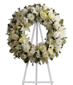 Funeral wreath with white roses, lilies, chrysanthemums, and green foliage on a white metal easel.